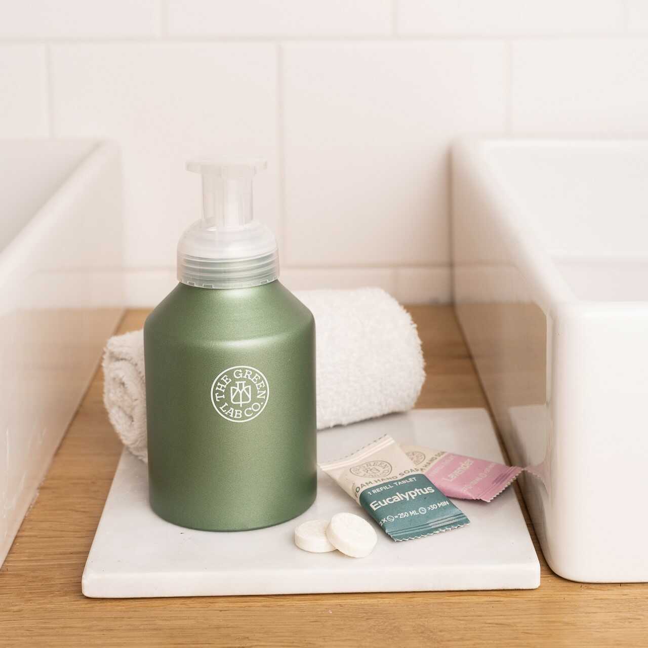 The Green Lab Co aluminum foaming soap dispenser on a white tray, shown with two white refill tablets and Eucalyptus/Lavender sachets beside a rolled towel between two sinks.